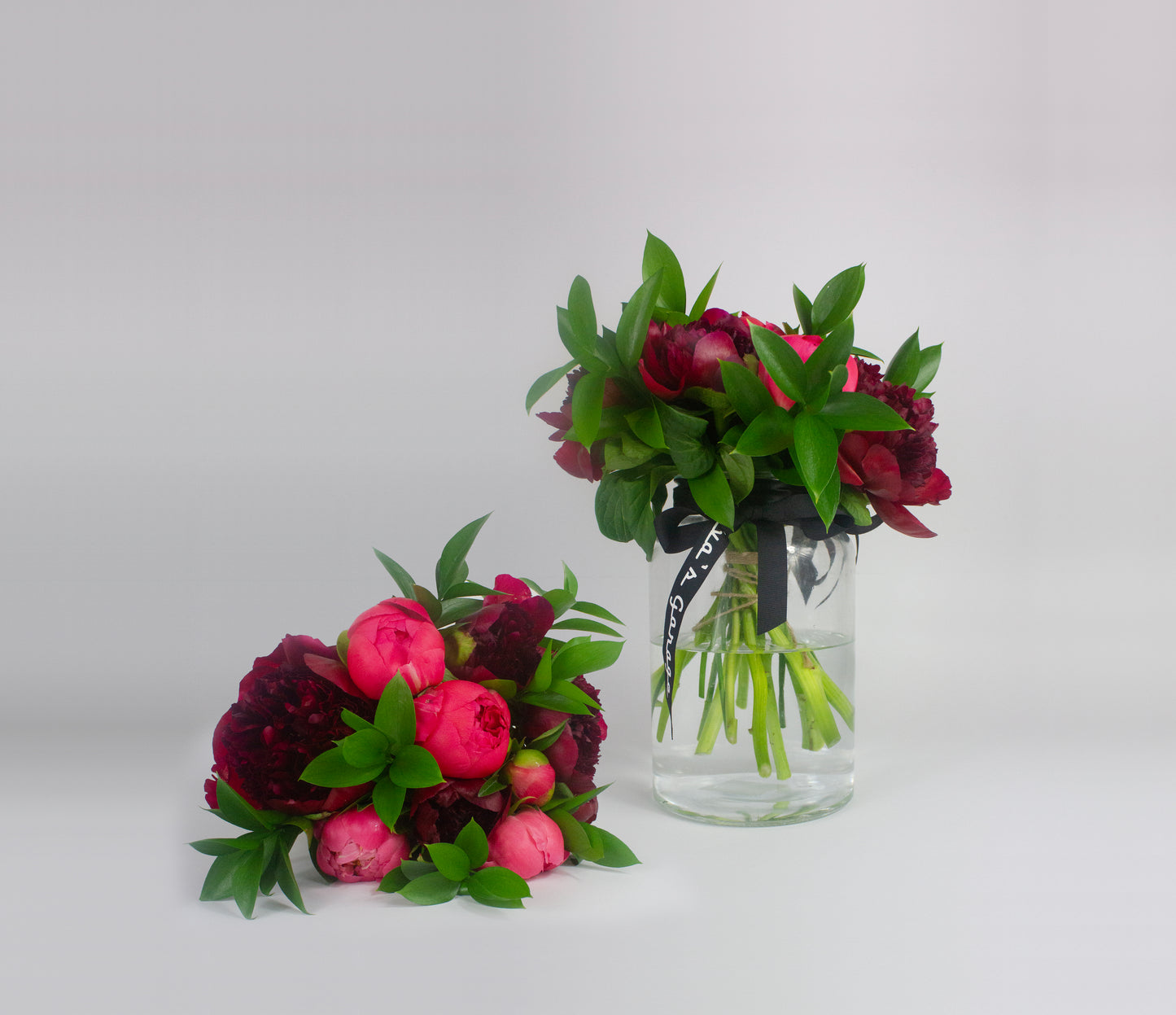 Two bouquets of red and pink Peonies with green leaves in a clear glass vase on a light gray background.