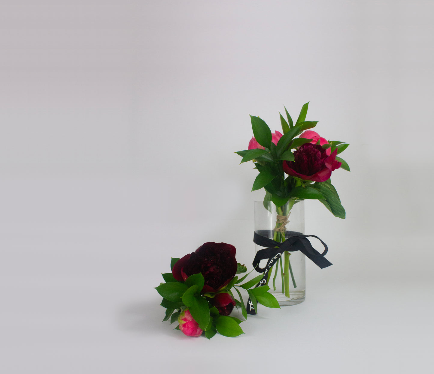 Two small floral arrangements in glass bottles with greenery and dark red flowers on a light gray background.