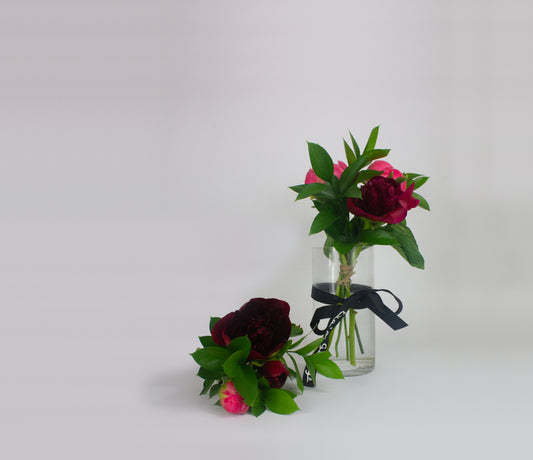 Two small floral arrangements in glass bottles with greenery and dark red flowers on a light gray background.